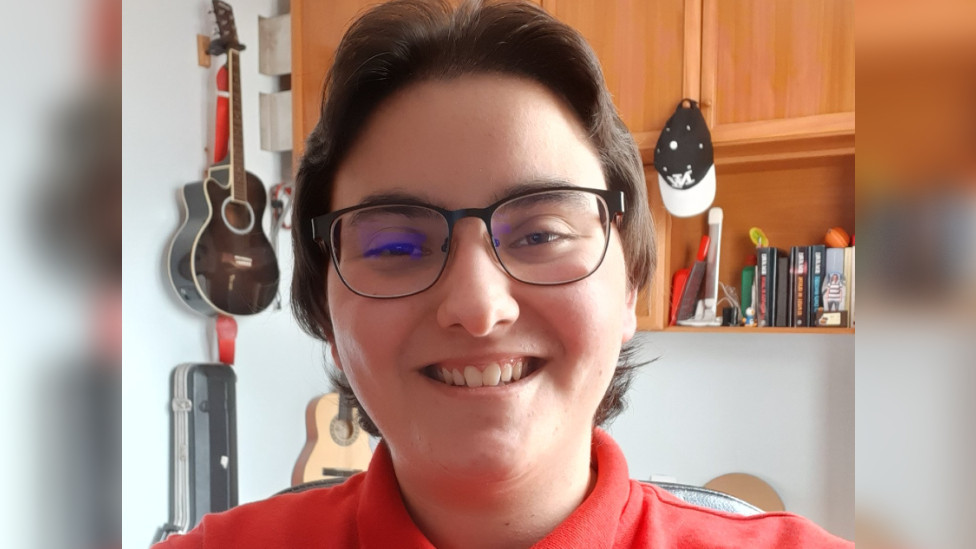 Daniel smiles at the camera. They are wearing glasses and a red top.