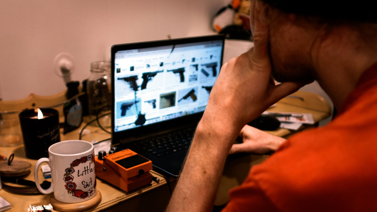 Max looking at a laptop with photos of handguns visible on the screen