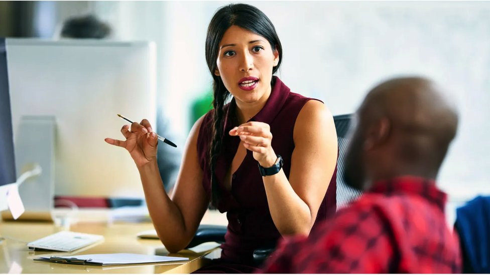 Woman at her desk using her hands to express what she is saying to a colleague