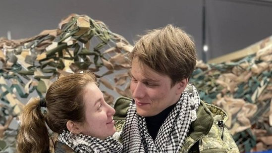 Yaryna Arieva and Svyatoslav Fursin hugging in military uniforms with a Ukrainian flag behind them