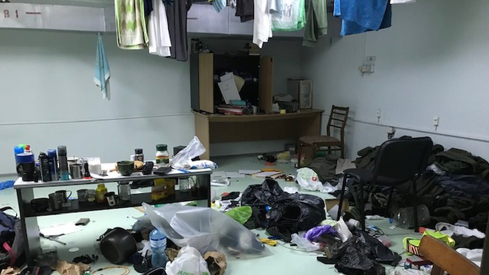 A chaotic room with a pale green floor covered with items like clothes, mugs, chairs, plastic bags