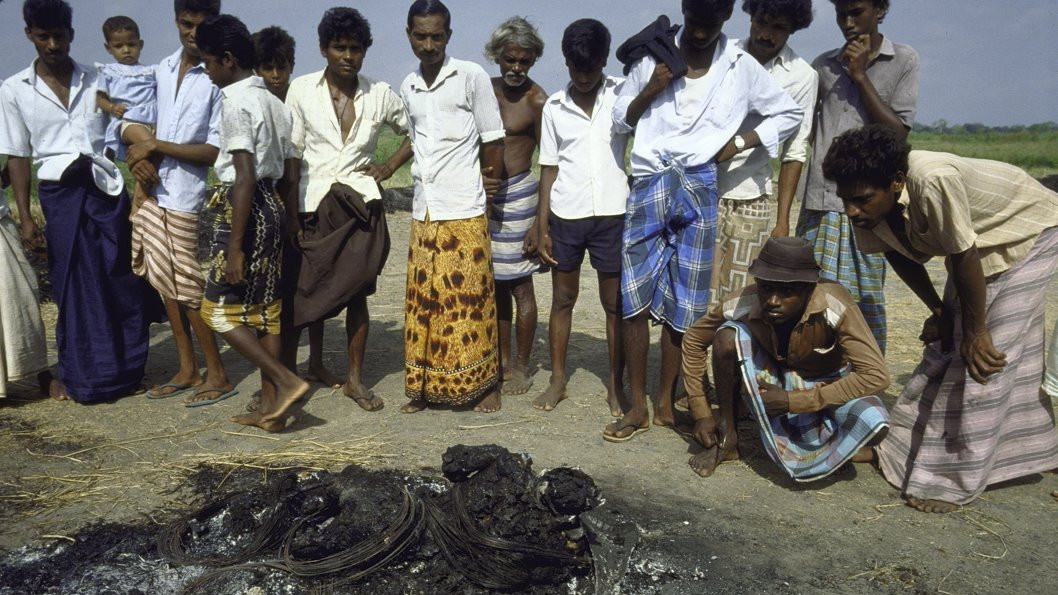 Burnt tyres lying on the ground with unidentifiable remains among them, while a group of men look on