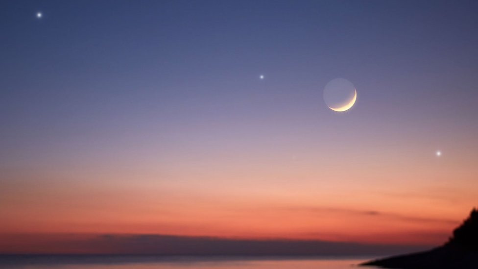 A crescent moon and three other illuminated bodies form a line above a mass of water and a cliff