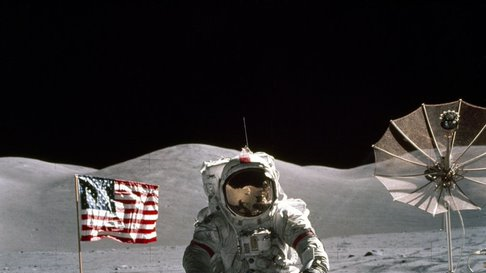 Apollo 17 Astronaut standing upon the lunar surface with the United States flag and a radio dish in the background