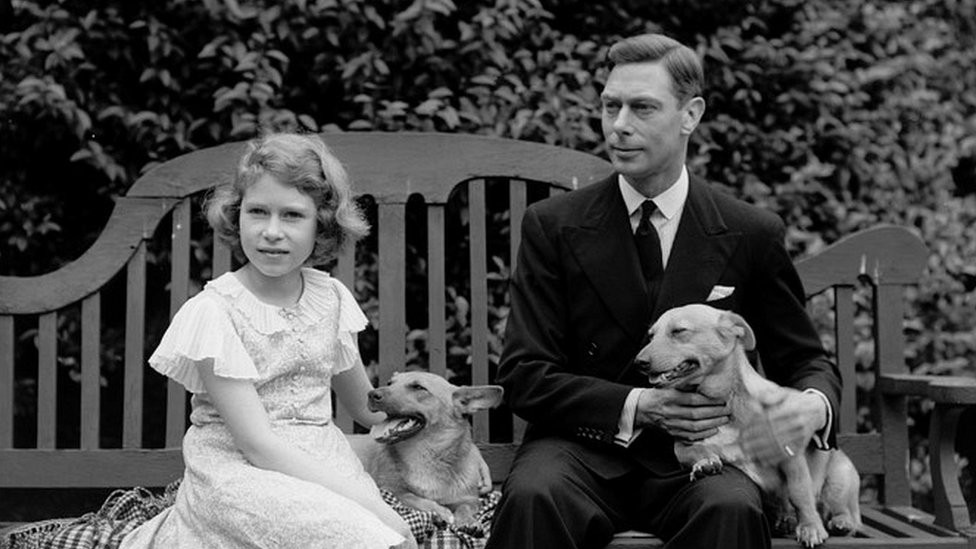 July 1936: George, Duke of York and Princess Elizabeth sitting on a bench with their corgi dogs in the grounds of their London home, 145 Piccadilly.