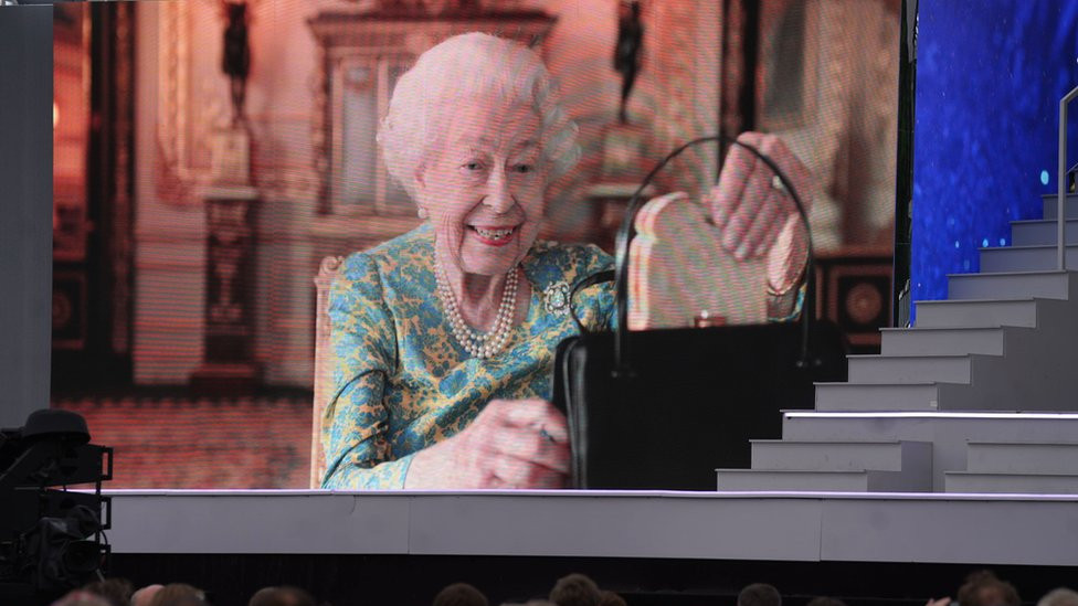 Kraljica vadi sendvič sa marmeladom iz ručne torbe, na velikom ekranu ispred Bakingemske palate