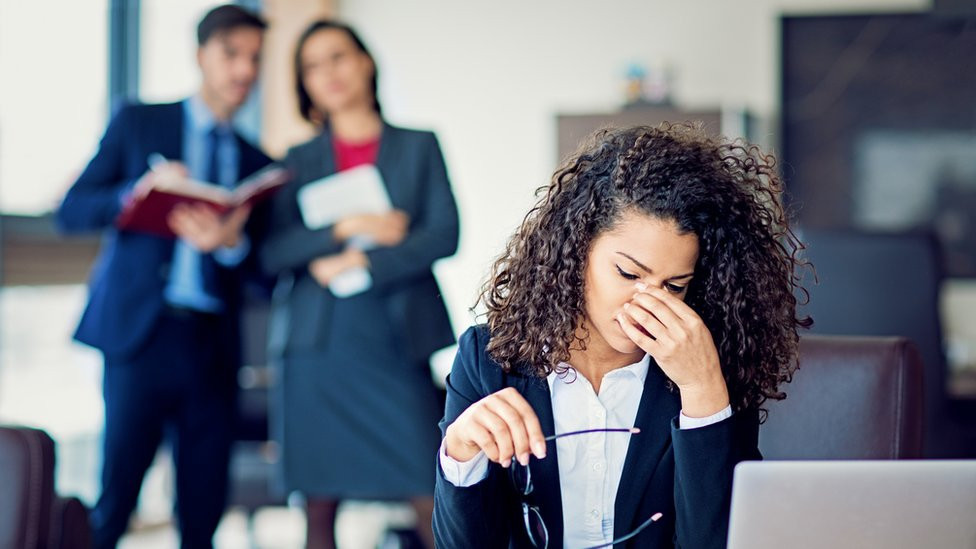 A young woman in an office holds her face in her hands as two colleagues talk behind her back