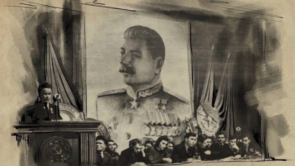 People sit at a desk under an enormous painting of Stalin, one man speaks from a lectern