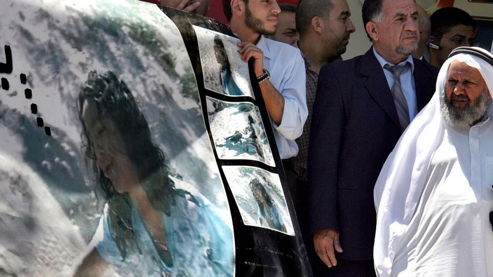 A pooster of Huda Ghalia reaching out to her fatyer's body while protesters hold the poster up