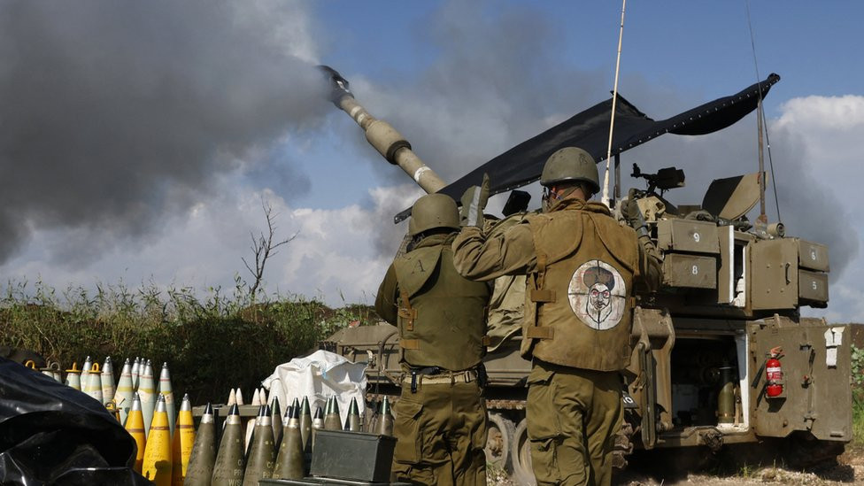 An Israeli soldier wearing a patch on the back of his flack jacket showing Hezbollah leader Hassan Nasrallah as a target, stands in front of a self-propelled artillery howitzer in northern Israel as it fires a shell towards southern Lebanon (4 January 2023)