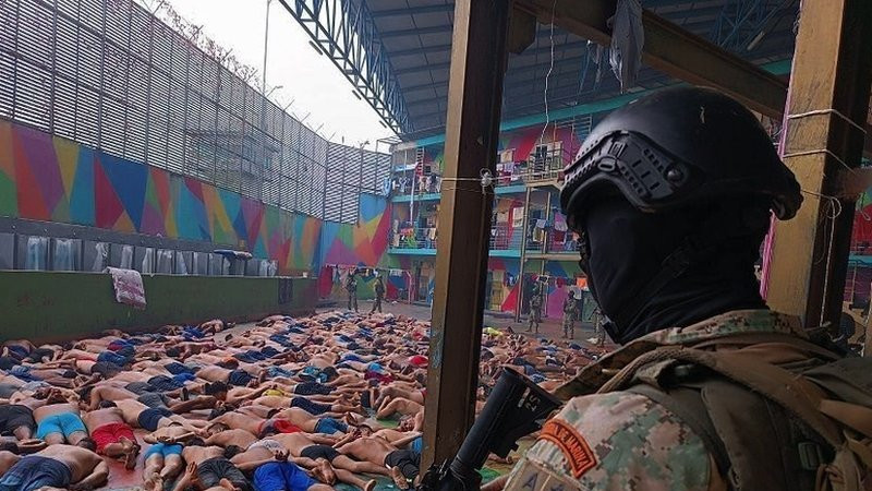 A soldier stands guard over dozens of prisoners, lying face down in a prison courtyard