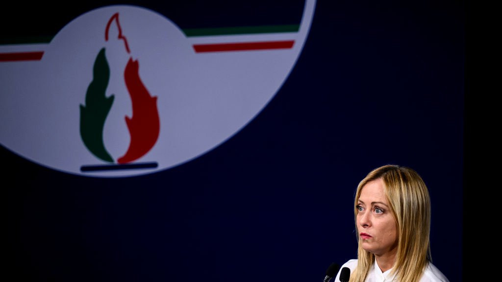 Giorgia Meloni standing next to a large sign of her party's logo, the tricolour flame