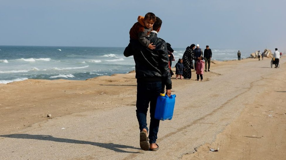 A man carries a child while holding a container, as displaced Palestinians, who fled their houses due to Israeli strikes, take shelter in a tent camp amid the ongoing conflict between Israel and Hamas, near the border with Egypt in Rafah in the southern Gaza Strip