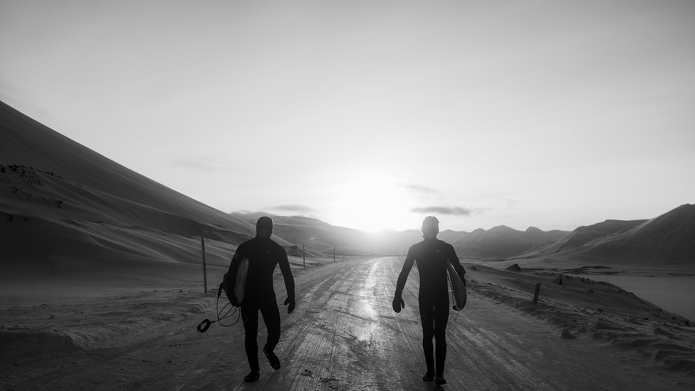 Surfers wearing wetsuits and holding surfboards walk along a path in Iceland