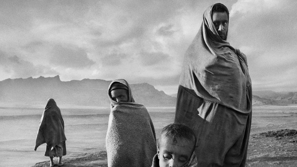 Draped in blankets to keep out the cold morning wind, refugees wait outside Korem camp. Ethiopia, 1984