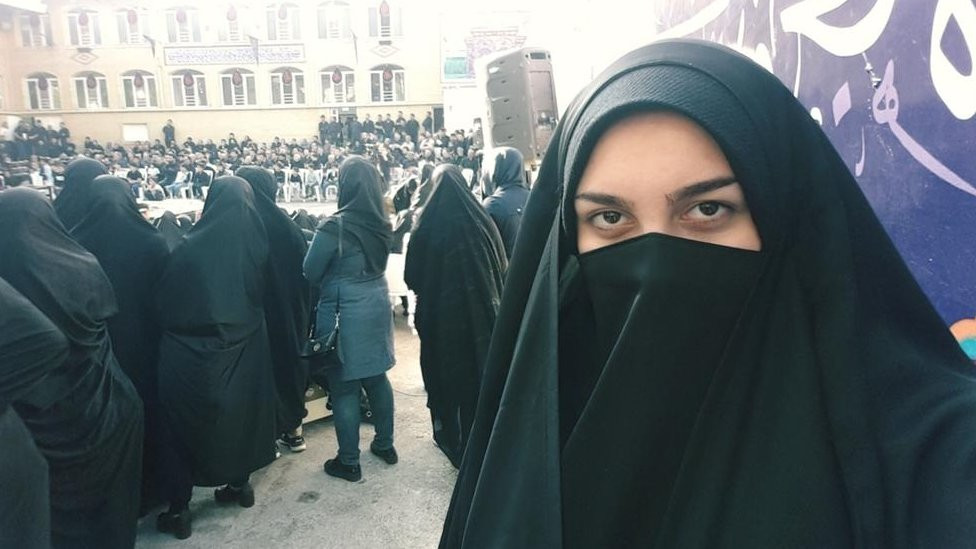 Ribell wearing a hijab that shows her eyes and forehead, standing in front of a lot of other people wearing a hijab