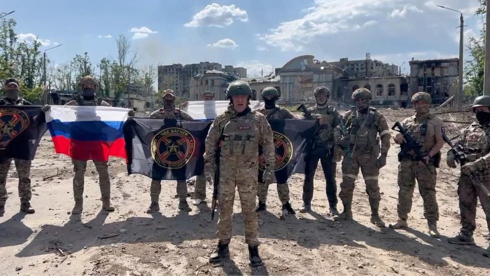 Prigožin u vojničkoj uniformi i sa puškom govori u videu o Bahmutu, dok iza njega stoje vojnici prekrivenih lica sa zastavama Rusije i Vagnera