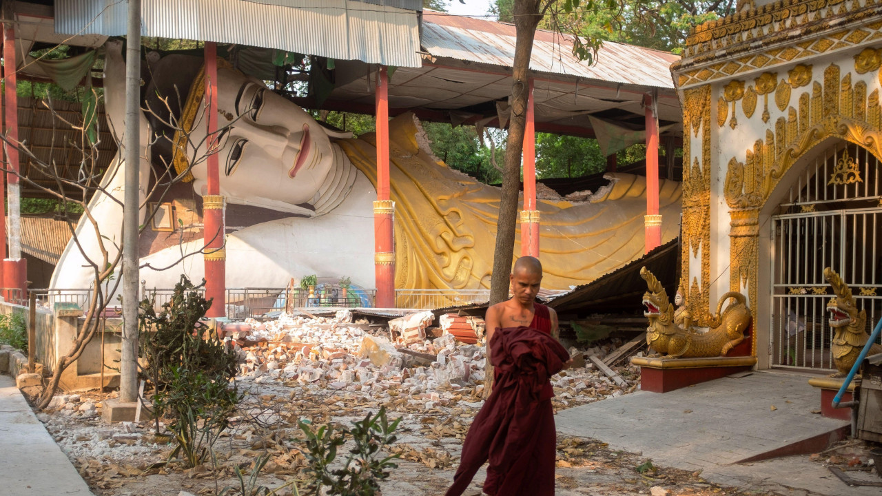 Srušena statua Bude i monah ispred koji hoda u crvenoj odori po ruševinama posle zemljotresa