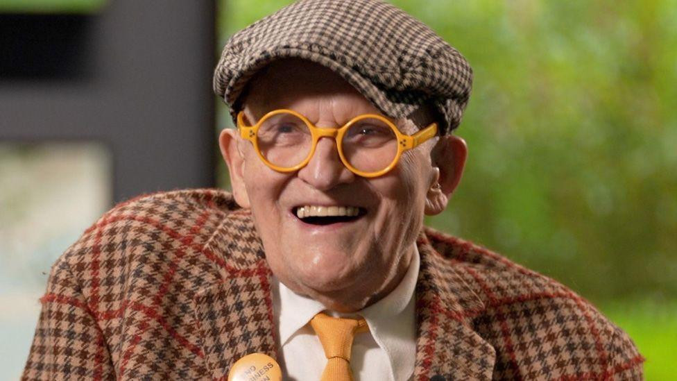 David Hockney smiling in yellow glasses in the Paris gallery