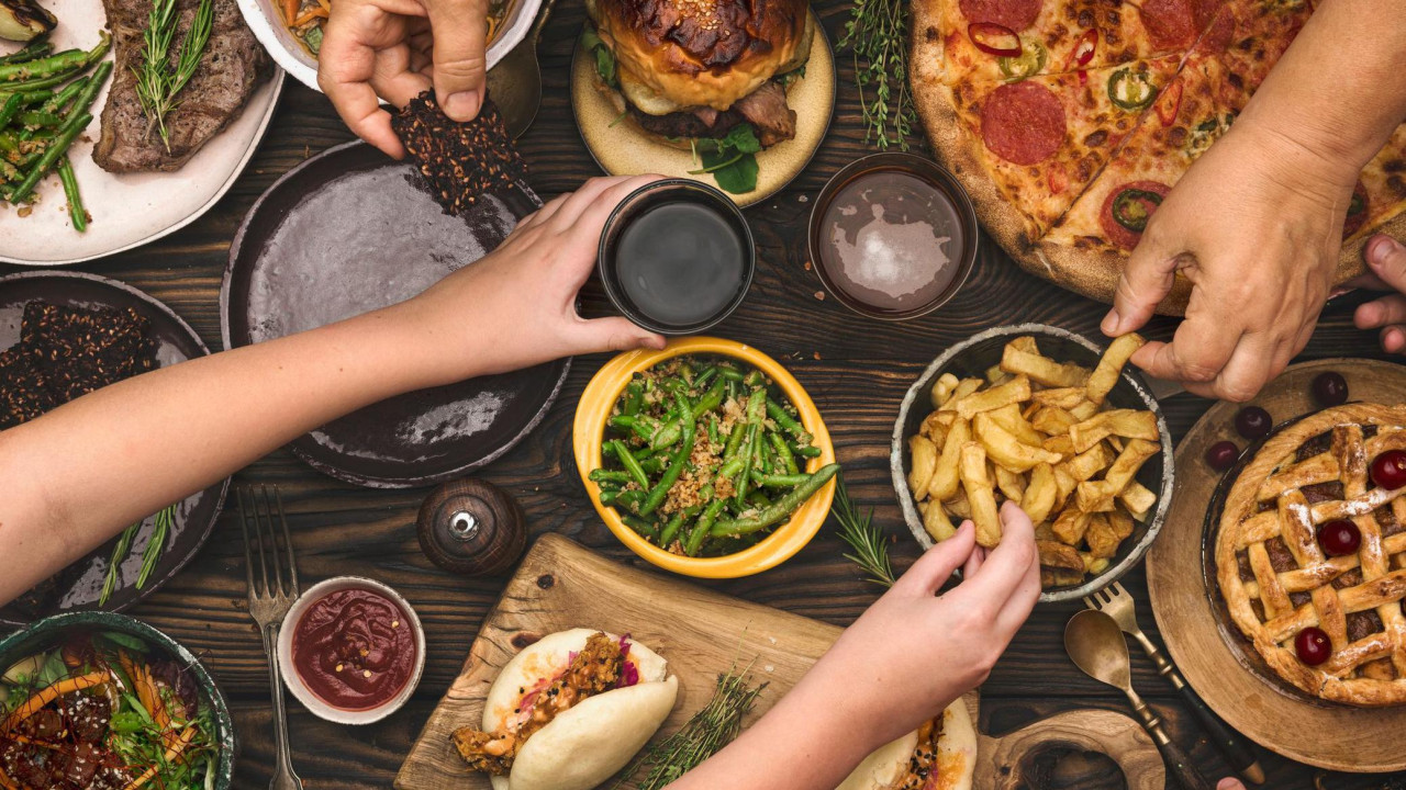 Ruke različitih ljudi biraju hranu, pomfrit, picu, salate i meso, iz činija na drvenom stolu