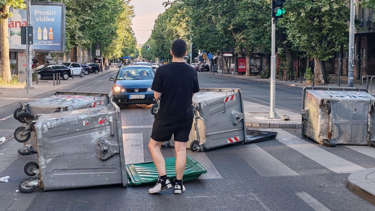 Momak okrenut leđima stoji na ulici ispred kontejnera i gleda u automobile