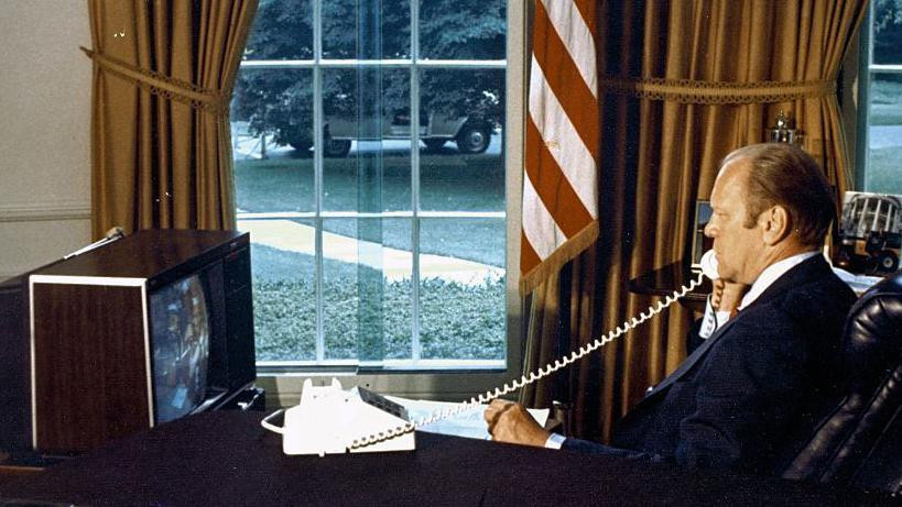 Američki predsednik Džerald Ford sedi za stolom u Ovalnom kabinetu i razgovara telefonom sa astronautima na brodu Apolo 1975. godine. Dok govori, gleda u monitor ispred sebe.