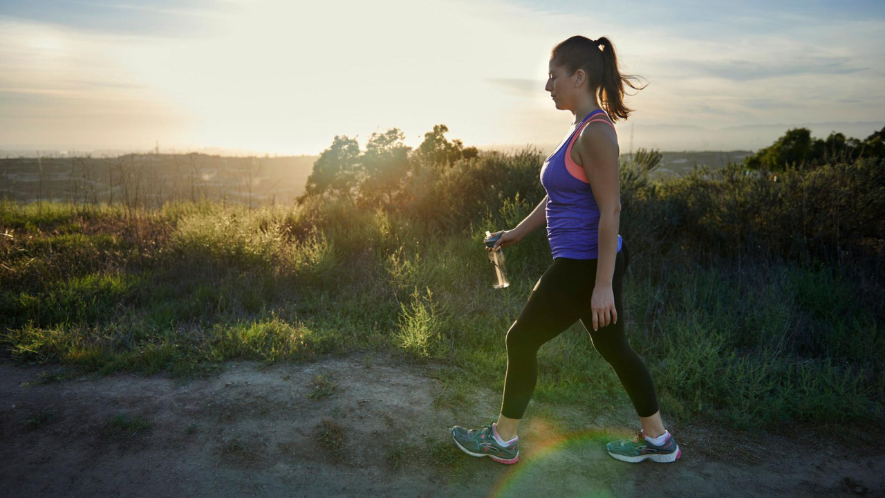 Žena u ljubičastoj majici i crnim helankama šeta selom. Kosa joj je vezana i drži flašicu vode.