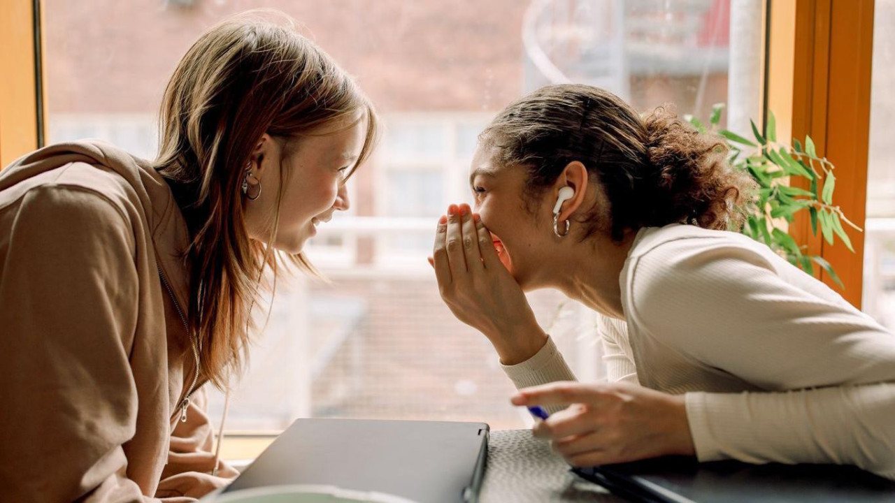 Dve mlade žene sede jedna preko puta druge i šapuću