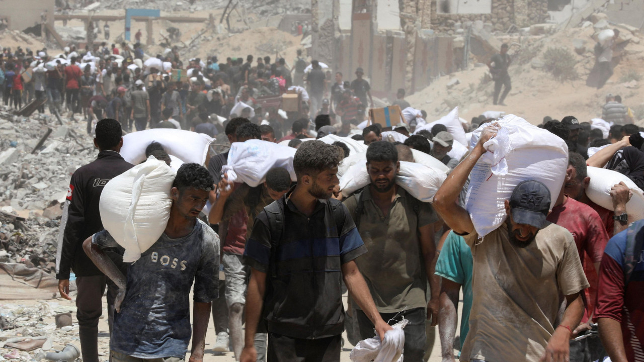 Ljudi na ramenima nose džakove dok je iza njih razrušen grad