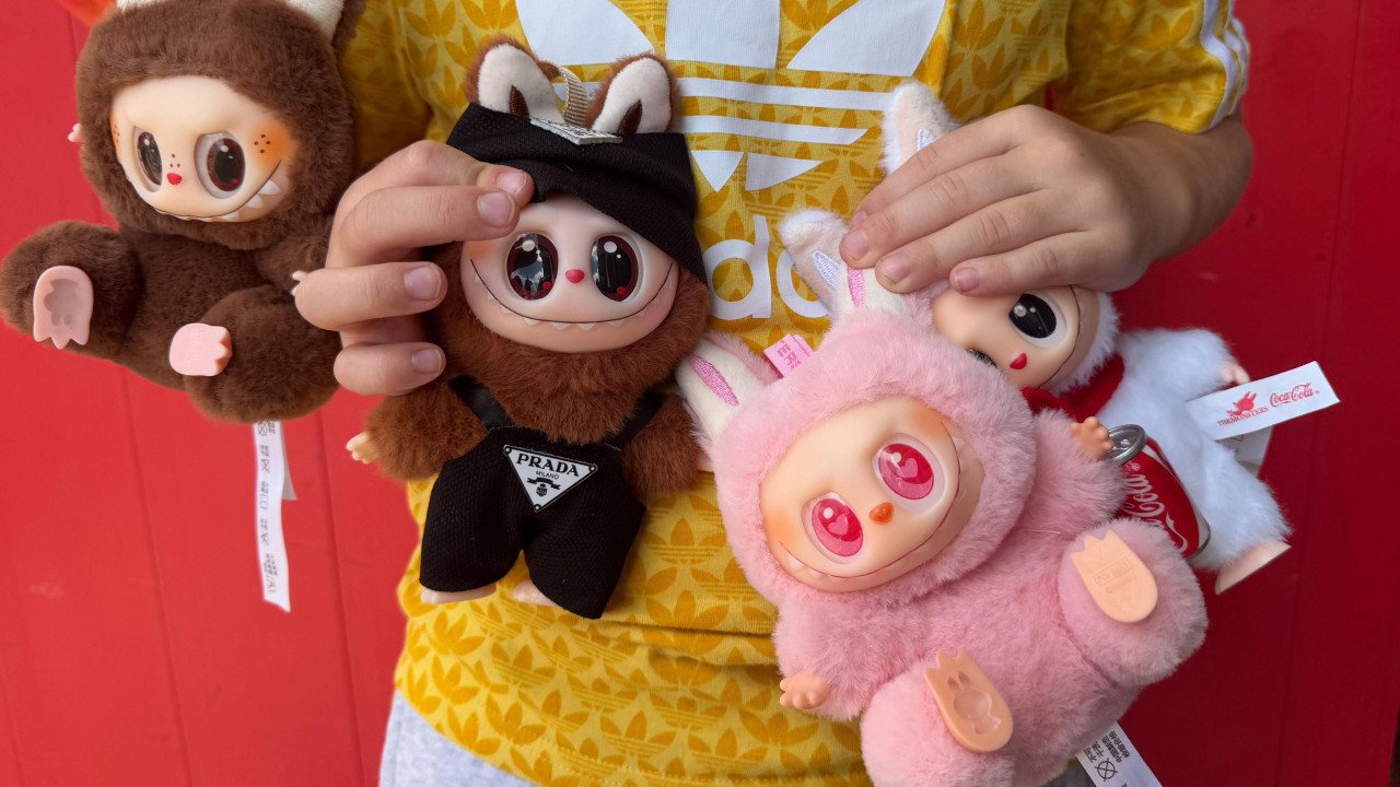 Dečak u žutoj sportskoj majici drži četiri lutke Labubu. Krznene lutke slične gremlinima imaju velike sjajne oči i osmehe sa širokim zubima. Ne vidi se Harijeva glava već njegove grudi, a fokus je na lutkama. Na levoj strani je jedna smeđa, druga smeđa sa crnim Prada kombinezonom, pored nje ružičasta, a na desnoj strani je bela koja drži limenku Koka-kole.