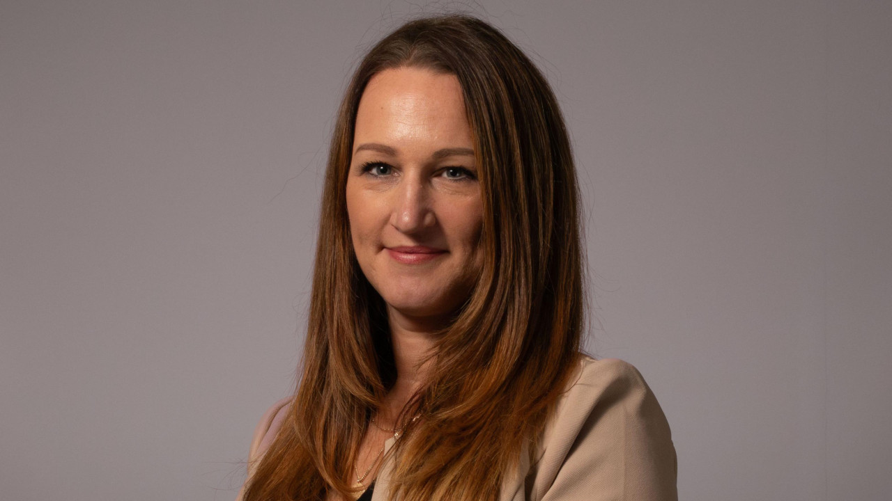 Kejt, elegantno obučena žena u bež odelu, sa dugom smeđom kosom, osmehuje se kameri.  