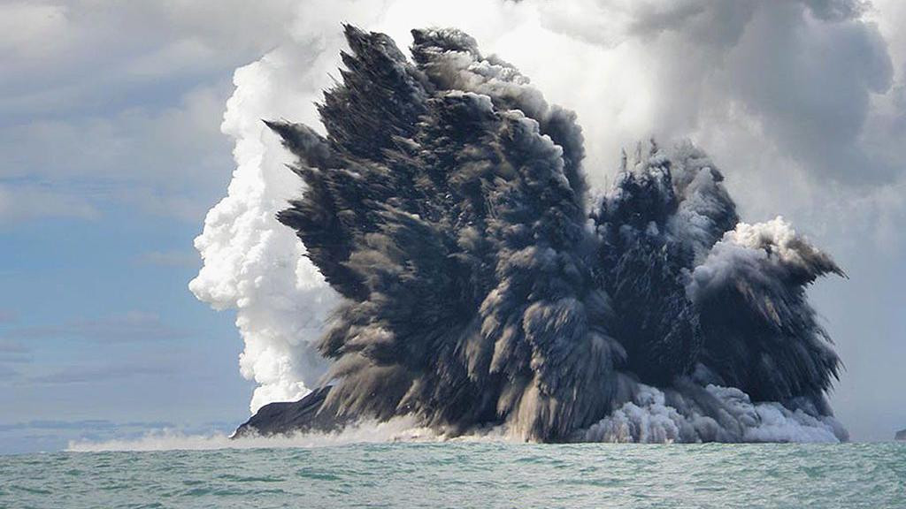 The photo shows the eruption of an underwater volcanoes. The eruption could be seen above sea level. A huge plume of black and white smoke is at the centre of the photo. The plume of smoke is coming from the water. Around the smoke there's just sea water and light blue sky.