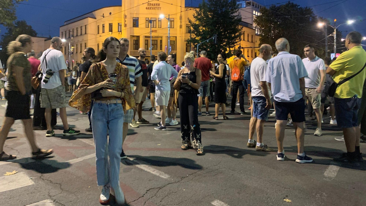 Devojka stoji na ulici među masom ispred Pranog fakulteta u beogradu, obučena je u farmerke i smeđu majicu, ima vezanu kosu i gleda u kameru, drži vodu u ruci