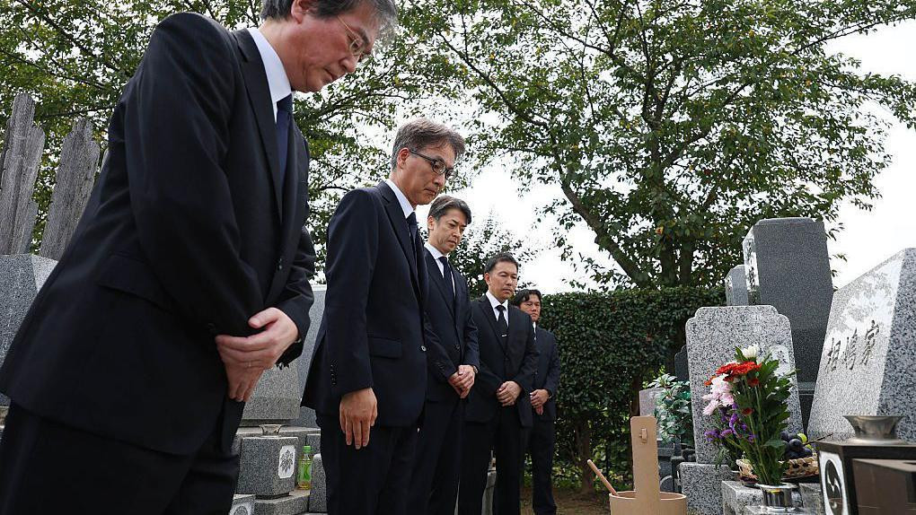 Pet muškaraca u tamnim odelima klanja se pred grobom u Japanu. Grob je napravljen od sivog mermera i ispred njega se nalazi vaza sa cvećem.