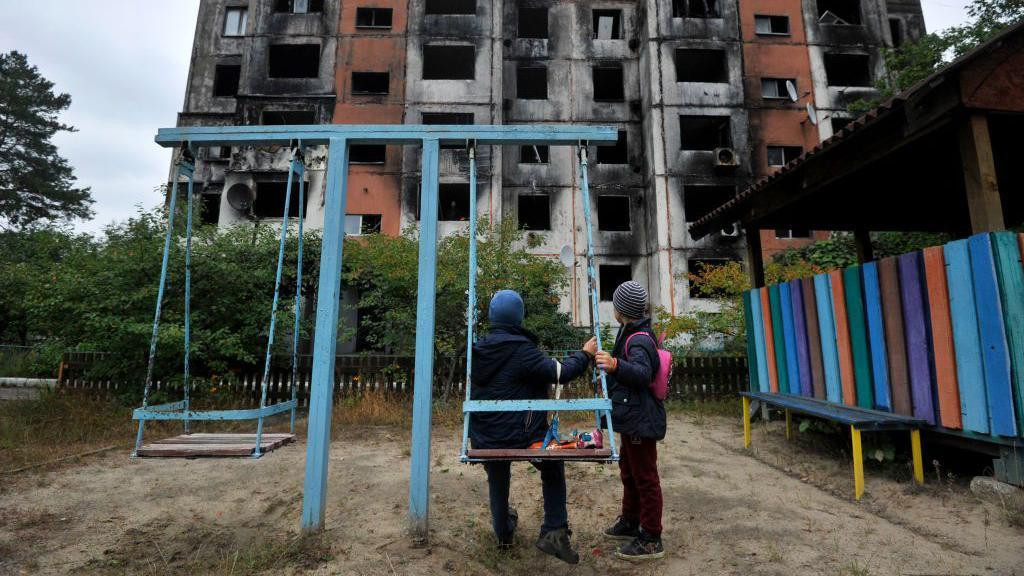 Deca na igralištu gledaju u spaljenu zgradu od bombardovanja 2022. godine