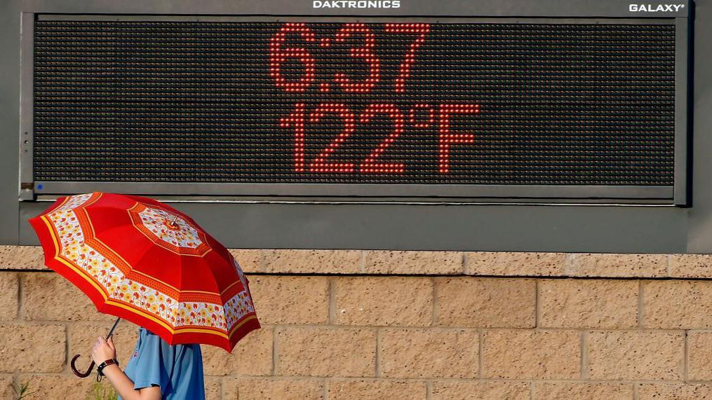 Osoba nosi kišobran umesto suncobrana prolazi pored oglasne table na kojoj je prikazana temperatura od 122 stepena Farenhajta