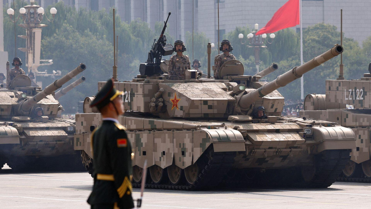 A soldier stands tall in the foreground while tanks carrying soldiers and guns pass during the parade, with a China flag behind.
