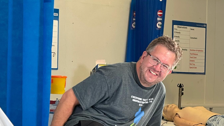 Nil Hoper sedi na ivici bolničkog kreveta, obučen u sivu majicu, crni šorc i naočare. Ne nosi proteze, tako da su njegove amputacije nogu vidljive. Smeje se u kameru.