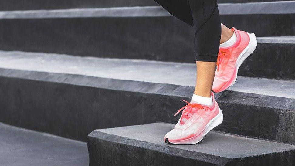 
                    A woman wearing black leggings and pink athletic shoes, standing on a set of black steps. One foot is placed on a higher step while the other remains on a lower one, sugg
