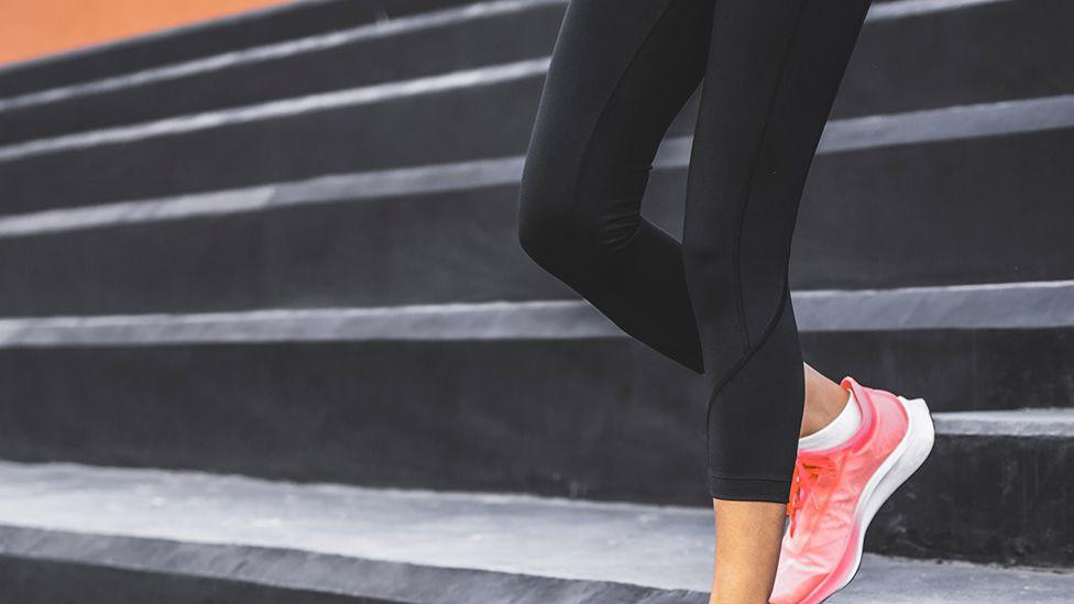 Žena u crnim helankama i ružičastim sportskim patikama stoji na crnim stepenicama. Jedno stopalo je na višoj stepenici, dok je drugo na nižoj