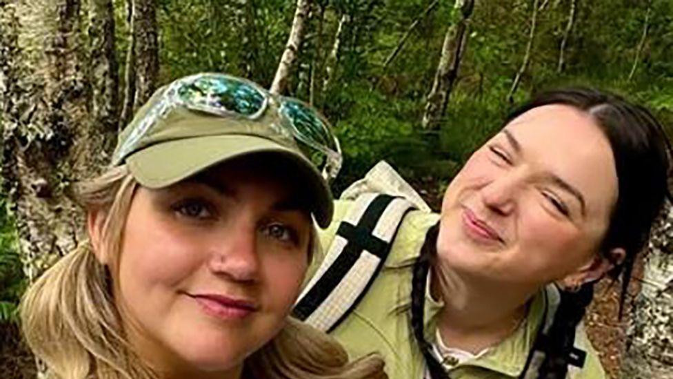 Emili i Lusi - dve žene stoje, okružene drvećem. Jedna nosi svetloplavu majicu i ima dugu plavu kosu. Druga je obučena u zelenu majicu i nosi ranac.