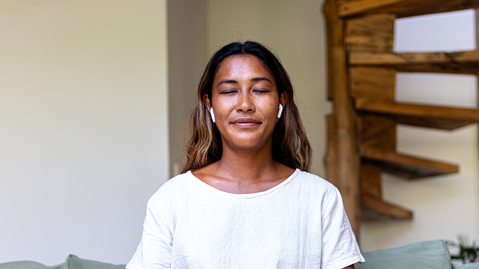 Žena sedi prekrštenih nogu na zelenoj sofi. Nosi belu majicu, sive pantalone i bežične slušalice. U pozadini se vidi drveno spiralno stepenište.