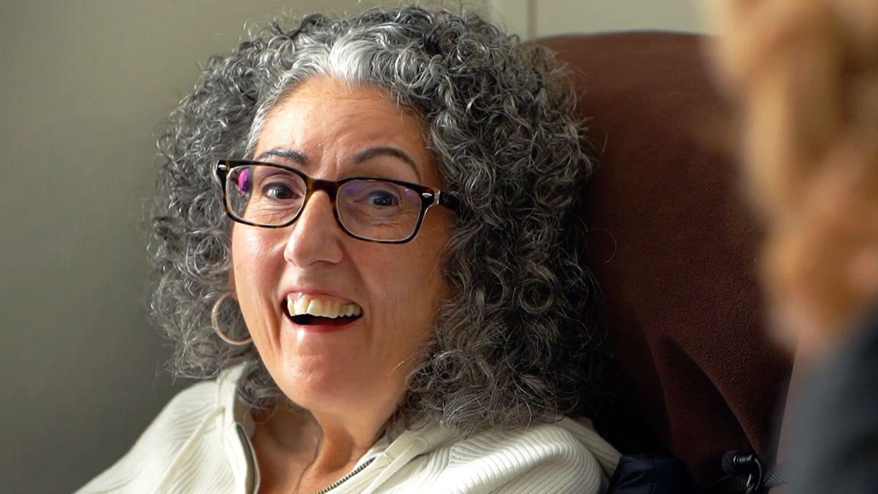 Sarah Ezekiel smiling. She is a woman with greying, curly, shoulder-length hair. She is wearing black-rimmed glasses.