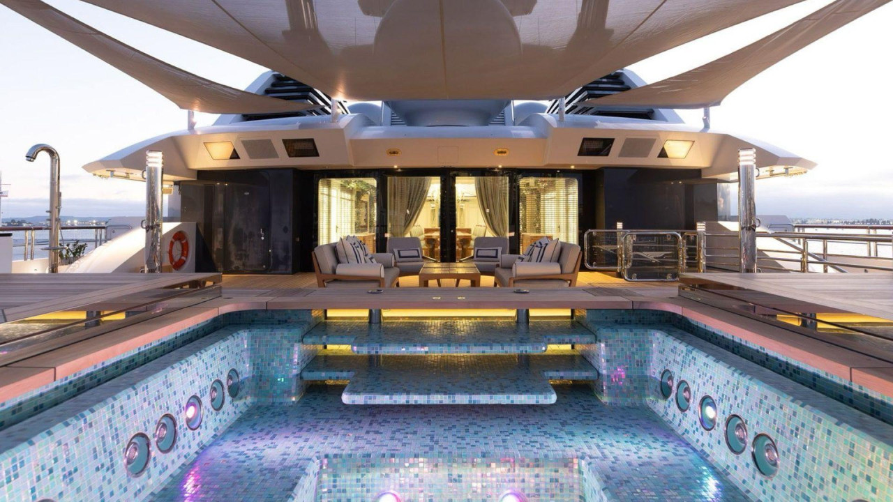 
                    The swimming pool on Amadea’s yacht, featuring small dark and light blue tiles. The pool is empty. There is a cover at the top of the photo and in the background a room i