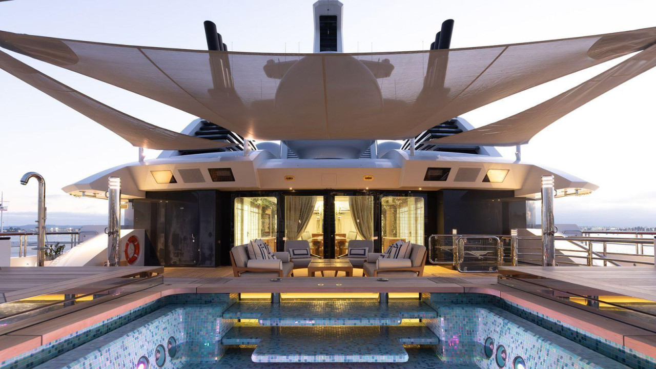 Bazen na jahti Amadea, sa malim tamnoplavim i svetloplavim pločicama. Bazen je prazan. Na vrhu fotografije je veliki suncobran iznad bazena, a u pozadini je osvetljena soba sa zavesama otvorenim preko prozora. Fotelje se nalaze na vrhu, proširenom delu pored bazena.