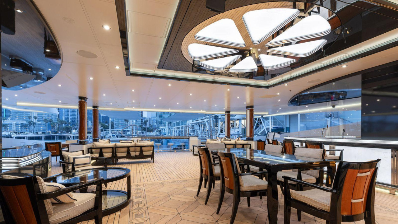 Soba unutar superjahte Amadea ima drvene podove i stolice sa bež jastucima, zajedno sa odgovarajućim sofama. Gradski pejzaž se vidi kroz prozore u pozadini.