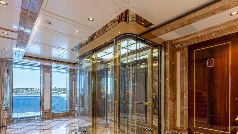 Lift sa zlatnim okvirom i staklenim zidovima, smešten uz zid. Pod je beo i sjajan sa pravougaonim smeđim ivicama. More i obala se vide kroz prozore u pozadini.