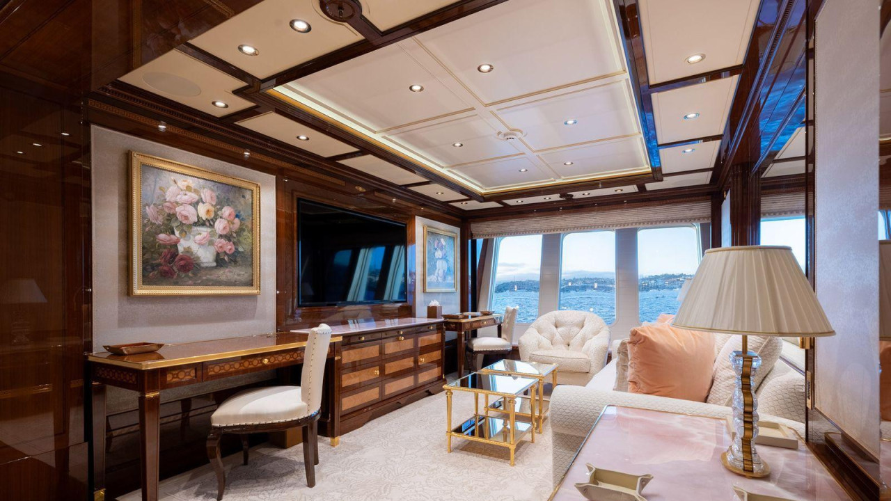 Jedna soba na superjahti Amadea opremljena je drvenim nameštajem, belim sofama i stolicom, televizorom i dvema cvetnim slikama. More i kopno se vide kroz prozore u pozadini.