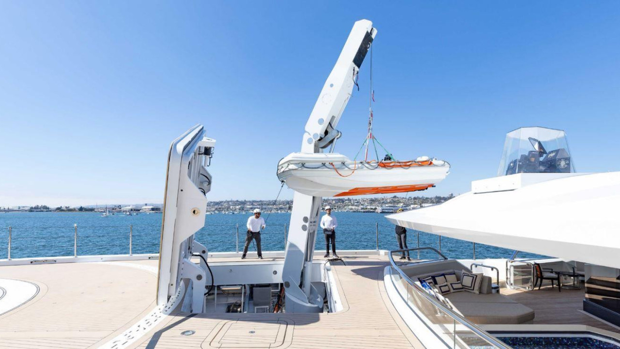 Dva muškarca u mornarskim uniformama stoje na palubi, sa čamcem podignutim iznad palube. Ivica helidroma je u levom delu fotografije, a desno je prostor za sedenje sa belim pokrivačem. Sunčan je dan, vidi se kopno u daljini. 