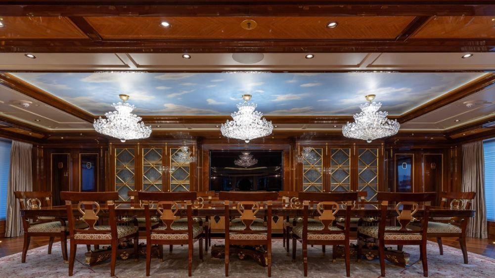 Drvene stolice poredane oko velikog drvenog stola u prostoriji sa tepihom i smeđim regalima okolo